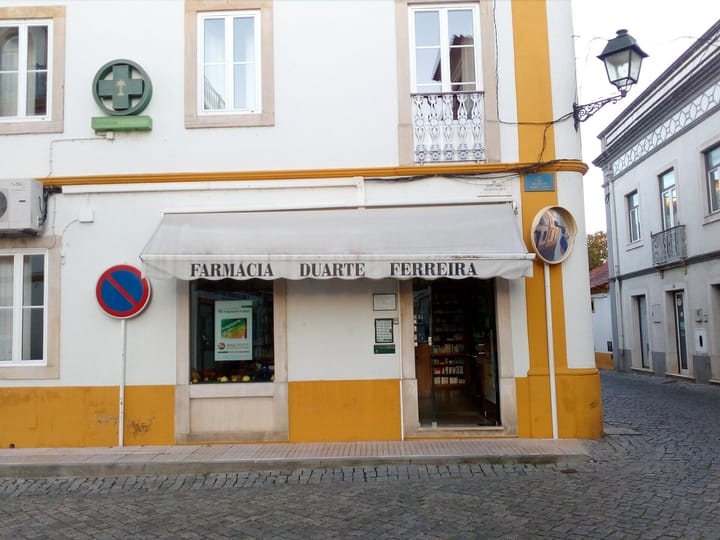 Farmacia Duarte Ferreira in Rossio ao Sul do Tejo, Abrantes, a corner pharmacy with a white awning, a green pharmacy cross on the yellow and white facade and calcada paving outside.