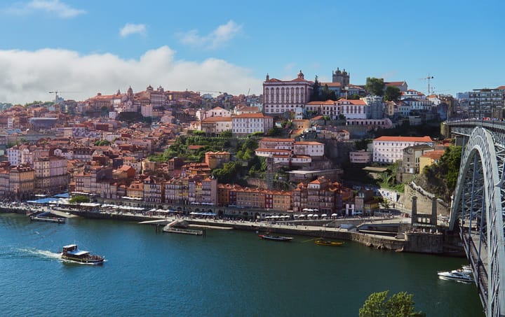 The Ribeira waterfront in Porto seen across the Douro on a clear day, with the steel arch of the Ponte Luis I at right and rabelo boats moored along the quay.