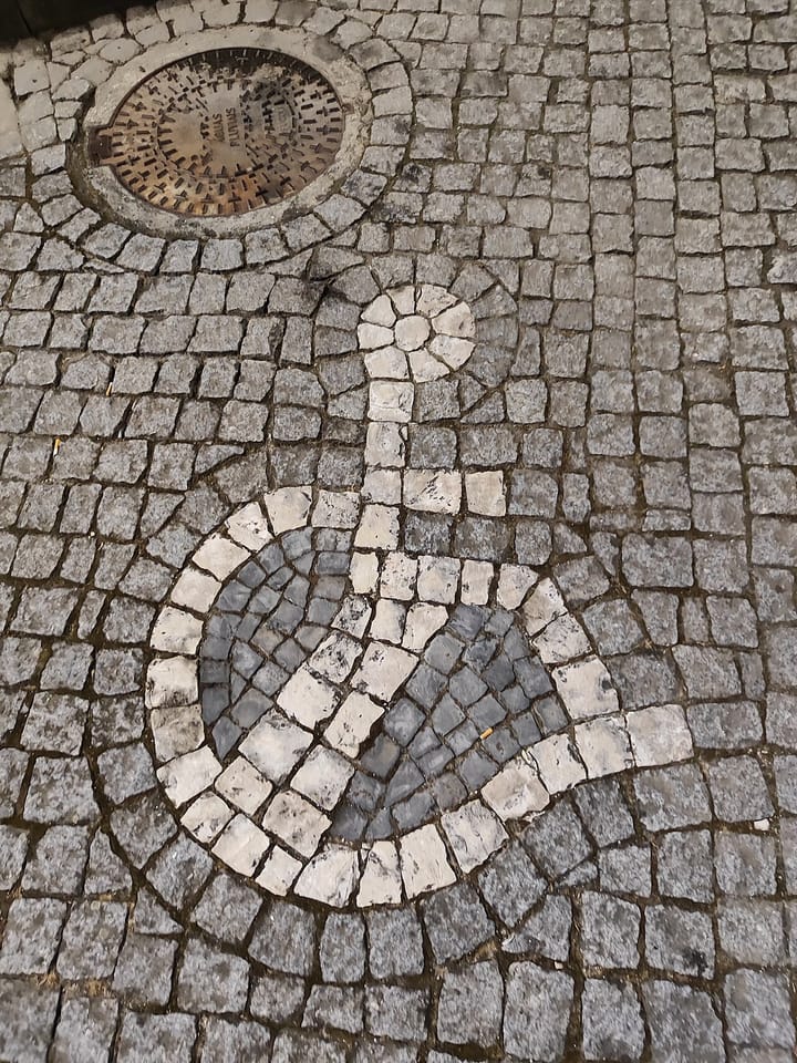 A wheelchair accessibility symbol laid in white and grey stones in traditional Portuguese calçada cobblestone pavement, beside a cast-iron drain cover