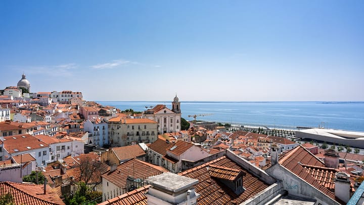 Terracotta rooftops of the Alfama district in Lisbon seen from a miradouro, with a church tower in the middle distance and the wide blue Tagus estuary beyond