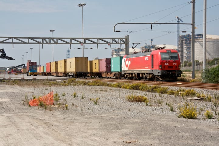 A red CP freight locomotive hauls shipping containers past the LNG tanks at Terminal XXI, Port of Sines.