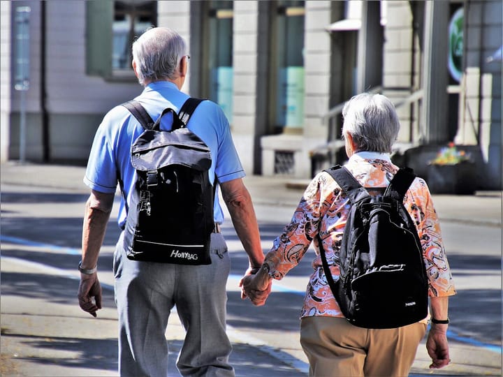 An elderly couple seen from behind walking hand in hand along a city street on a sunny day