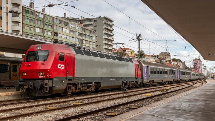 A red and grey CP locomotive at the head of a rake of stainless-steel Sorefame Intercidades carriages under the platform canopy at Lisbon's Santa Apolonia station