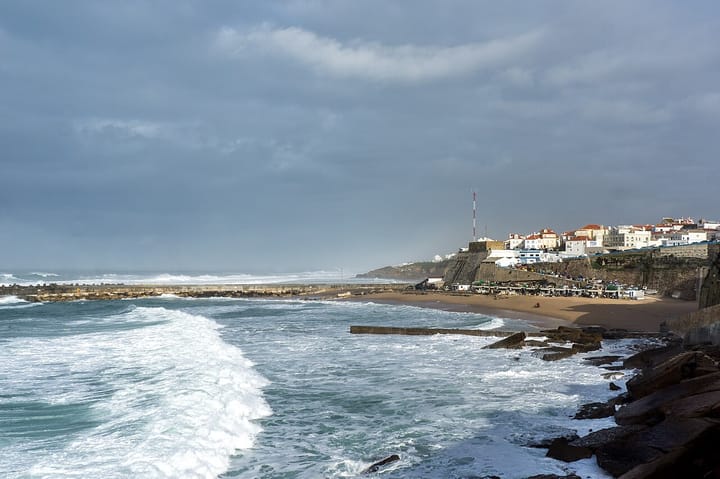 The whitewashed houses of Ericeira on the clifftop above the fishing beach, with Atlantic surf breaking over the rocks and breakwater below a grey sky
