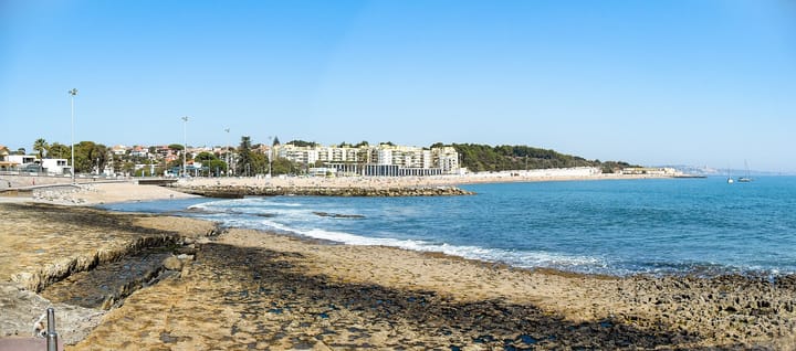 The seafront at Santo Amaro de Oeiras west of Lisbon, with the sandy bay, breakwater rocks and apartment blocks behind under a clear blue sky