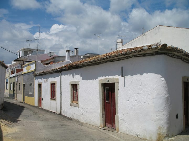 A street of low whitewashed houses with terracotta pantile roofs and red doors in Sao Bras de Alportel, in the Algarve, under a blue sky with cumulus cloud