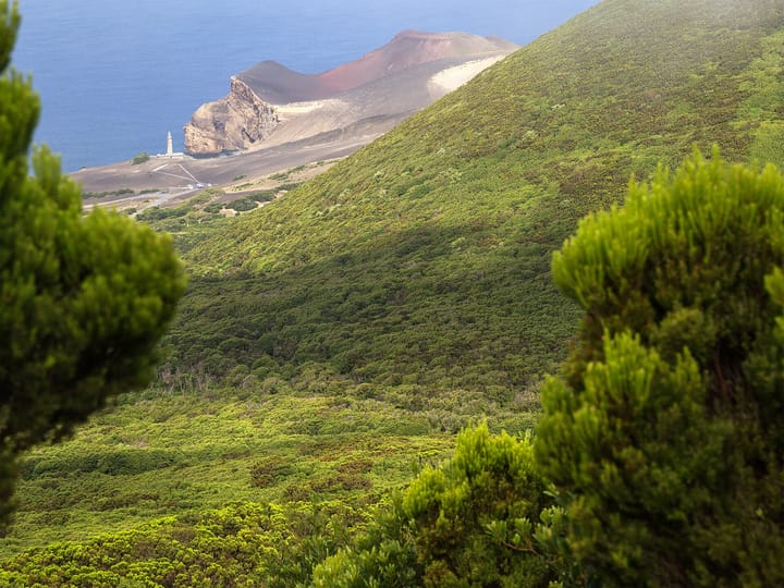 The Capelinhos volcano cone and lighthouse on the coast of Faial island in the Azores, seen across green forested hillsides above the Atlantic Ocean