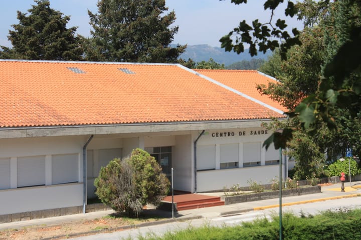 The low white building of the Melgaco health centre in northern Portugal, with a red-tiled roof and CENTRO DE SAUDE lettering above the entrance, wooded hills rising behind it.