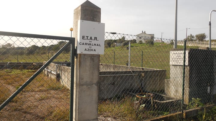 The gate of the ETAR de Carvalhal de Azoia, a small municipal wastewater treatment plant in the Soure countryside, with concrete tanks behind the fence and a farmhouse on the hill beyond.
