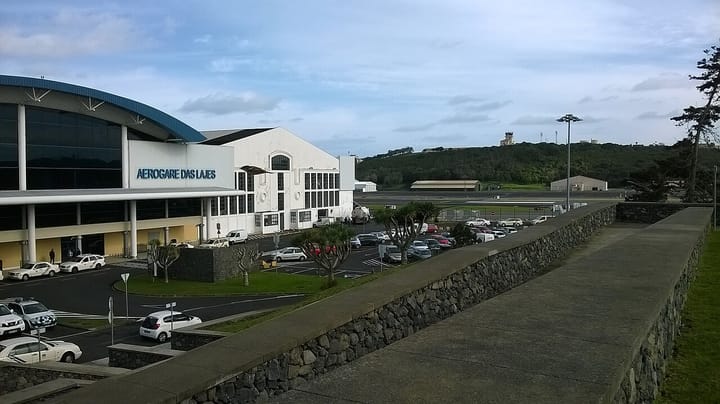 The Aerogare das Lajes terminal on Terceira in the Azores, a white building with a curved blue roof, seen across its car park with the airfield behind it under a cloudy sky.