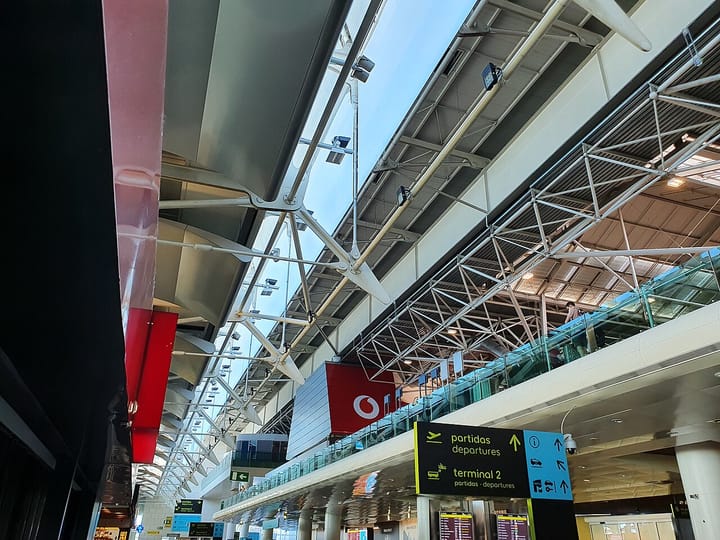 The white steel roof trusses spanning the departures level of Terminal 1 at Lisbon's Humberto Delgado airport, with yellow partidas and departures signage below.