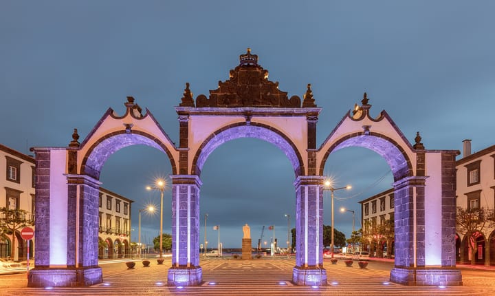 The three floodlit basalt arches of the Portas da Cidade in Ponta Delgada, Sao Miguel, at dusk, with the harbour and a statue framed through the central archway.