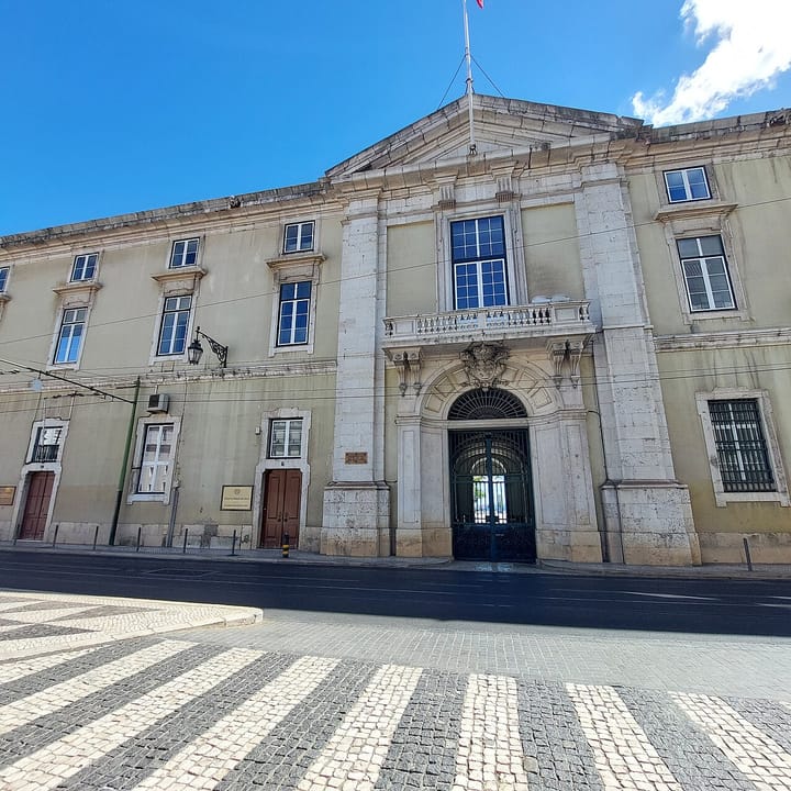 The neoclassical stone facade of the Tribunal da Relacao de Lisboa beside the Pacos do Concelho, with its arched iron gate and brass plaques above black-and-white calcada paving.
