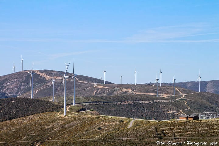 A row of white wind turbines spread across dry hills at the Pampilhosa da Serra wind park in central Portugal, with a substation building on the right