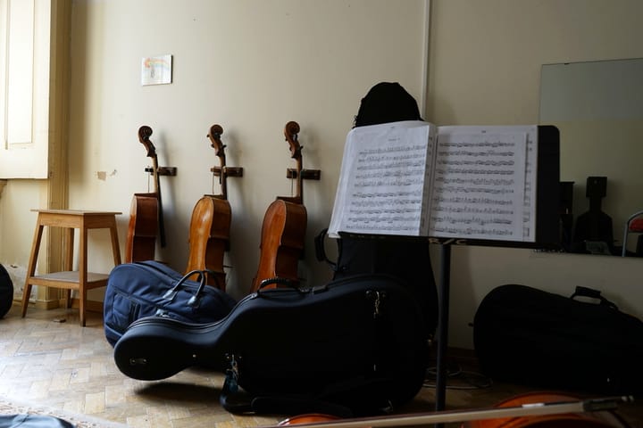 Three cellos on wall racks in a Portuguese music school classroom, with instrument cases on the parquet floor and open sheet music on a stand