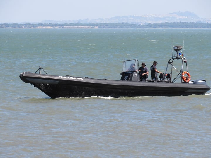 A black Policia Maritima rigid inflatable patrol boat under way on an estuary in Portugal with three officers aboard and a wooded shoreline behind