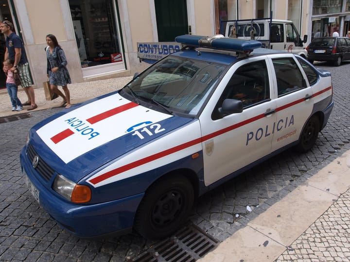 A Policia de Seguranca Publica patrol car in white and blue livery parked on a cobbled Portuguese street beside a pavement, with shopfronts and pedestrians behind
