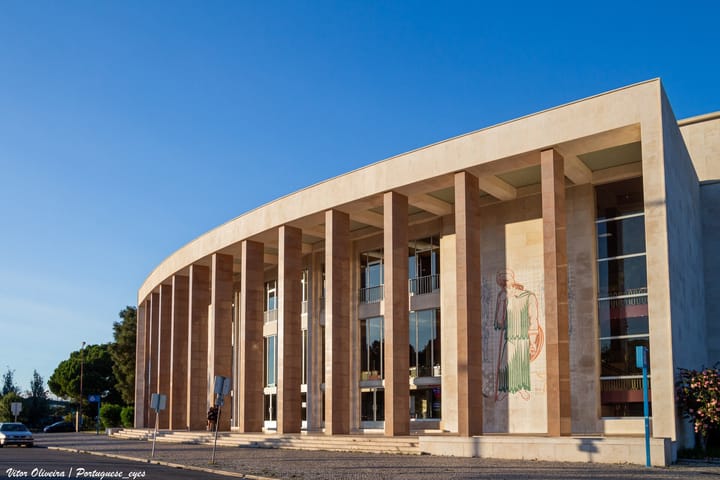 The curved colonnaded stone facade of the Aula Magna and rectory building of the Universidade de Lisboa under a clear blue sky, with a figurative mural on the end wall