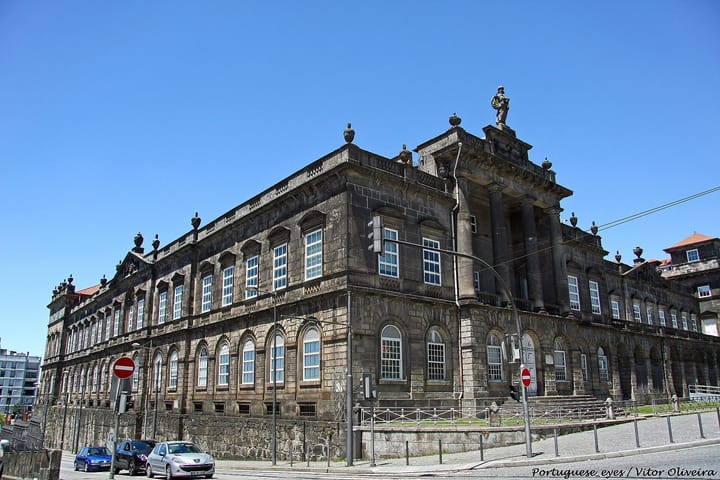 The neoclassical granite block of the Hospital de Santo Antonio in Porto seen from a street corner, its pediment topped by a stone statue against a clear sky