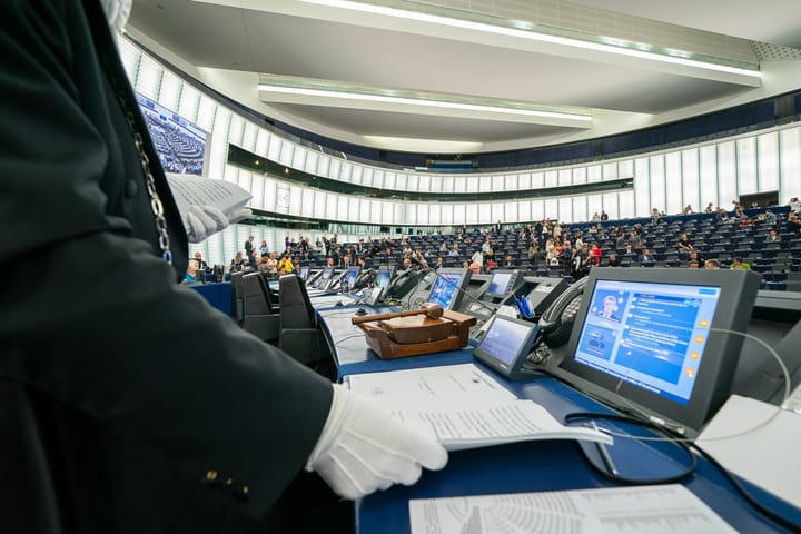 The European Parliament hemicycle in Strasbourg seen from the president's desk, a wooden gavel in the foreground and members taking their seats in the chamber beyond