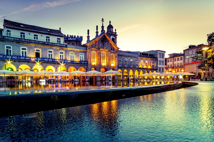 The Praca da Republica in Braga at dusk, with the lit baroque facade of the Igreja da Lapa above the arcaded Arcada and cafe umbrellas reflected in the water