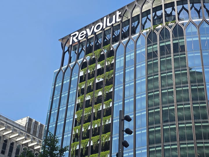 The glass and steel facade of Revolut's headquarters building seen from street level against a clear blue sky, the company name in white lettering along the roofline