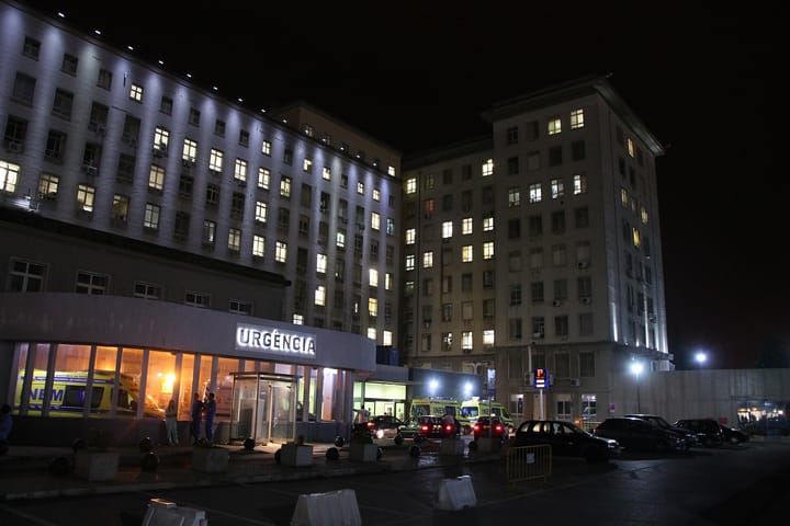 The lit Urgencia entrance of the Hospital de Santa Maria in Lisbon at night, with INEM ambulances parked outside and staff standing by the revolving door