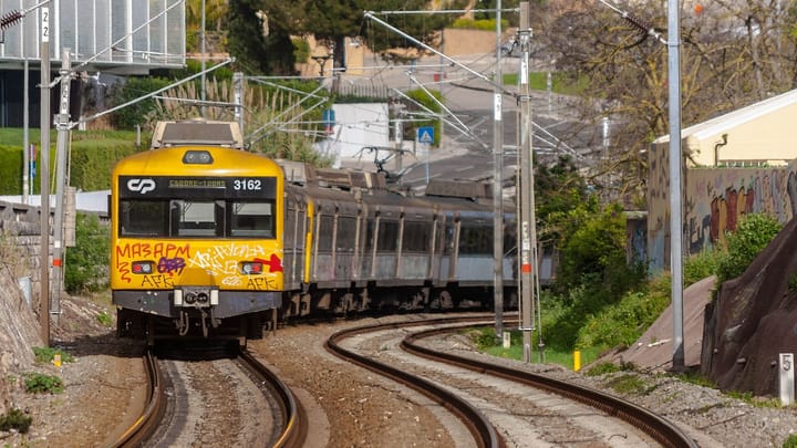 A yellow-fronted CP commuter train bound for Cais do Sodre rounds a curve on the Cascais line at Caxias, between trees and overhead wires
