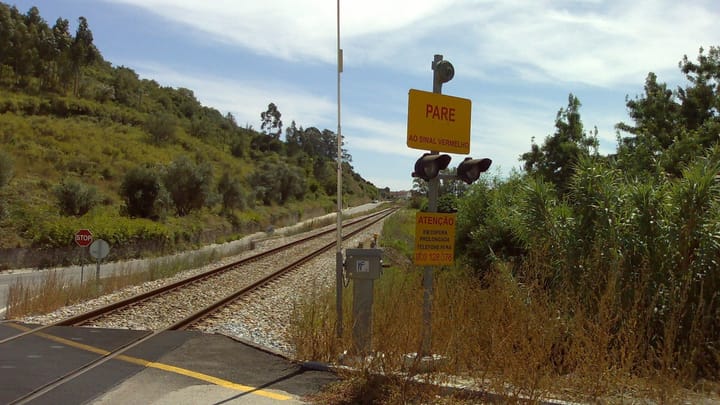 An automatic level crossing at Zibreira, with a yellow sign reading Pare ao sinal vermelho above two warning lights, and a single railway track running into green hills