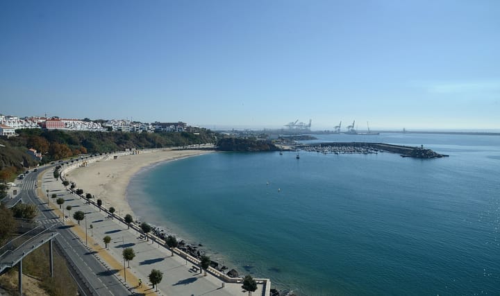 The bay of Sines seen from the old town, with a sandy beach and seafront road in the foreground, the marina breakwater, and the container cranes of the port on the horizon