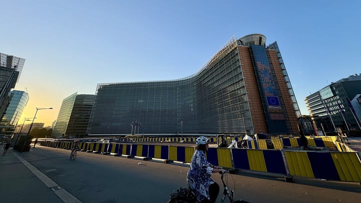 The curved glass facade of the Berlaymont, the European Commission's headquarters in Brussels, at sunset, with a cyclist passing blue and yellow barriers in front