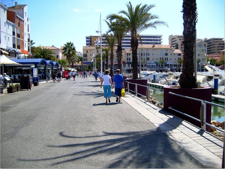 The pedestrian quay at Vilamoura marina in the Algarve, lined with palms and moored motor yachts, with holiday apartment blocks and hotel buildings rising behind the waterfront shops.