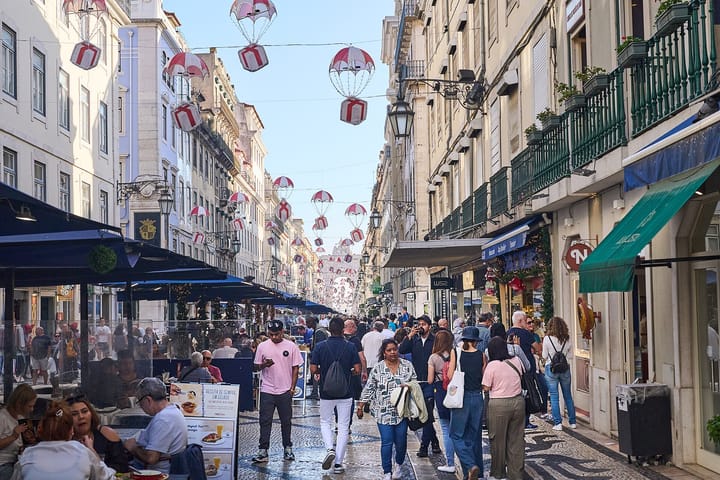 Rua Augusta in Lisbon, with restaurant esplanades under awnings on the left, a crowd on the mosaic pavement and a municipal waste bin at the right edge.