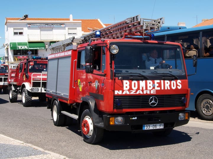 A red Mercedes fire appliance lettered BOMBEIROS NAZARÉ on a Portuguese street, ladders on the roof and two more Nazaré appliances parked behind it.