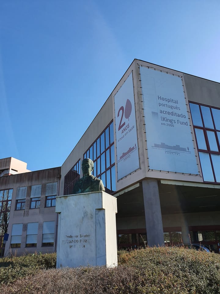 The Hospital Professor Doutor Fernando Fonseca in Amadora seen from below, with a bronze bust on a stone plinth in front of the entrance canopy.