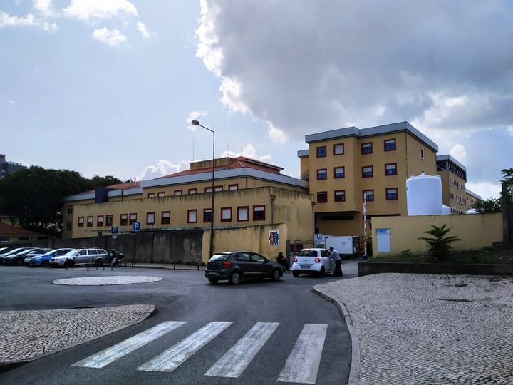 The Hospital Curry Cabral in Lisbon seen from the street, its yellow blocks with red window surrounds standing behind a low wall, with cars parked outside and a crossing in the foreground.