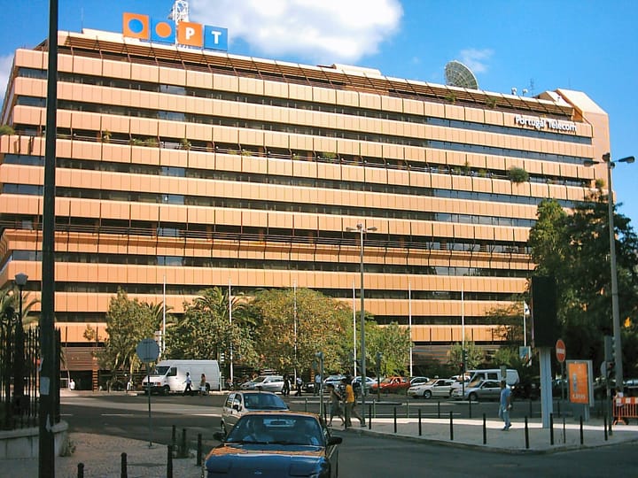 The Portugal Telecom head office in Lisbon, a long terraced facade with a rooftop PT sign and a satellite dish behind it, cars and pedestrians on the street below.