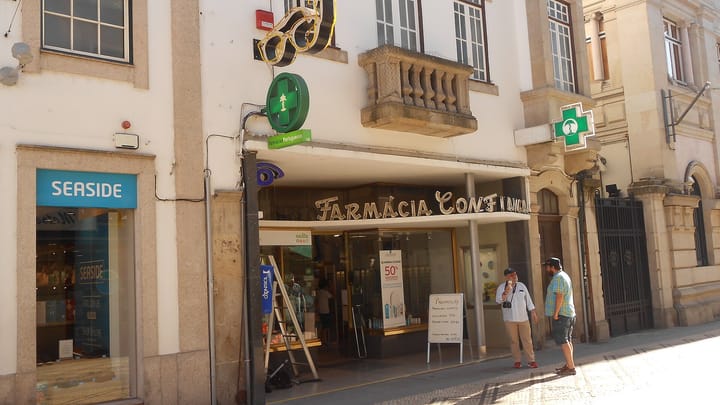 The shopfront of a working high-street pharmacy in Viseu, with the green pharmacy cross lit above the pavement and two people standing outside.