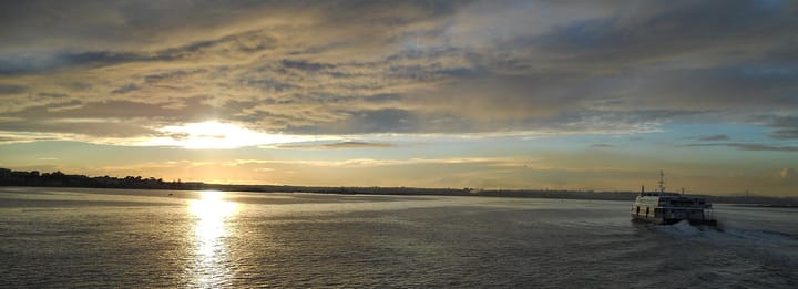 A Transtejo catamaran under way on the Tagus towards Lisbon in late afternoon light, with the 25 de Abril bridge on the horizon behind it.