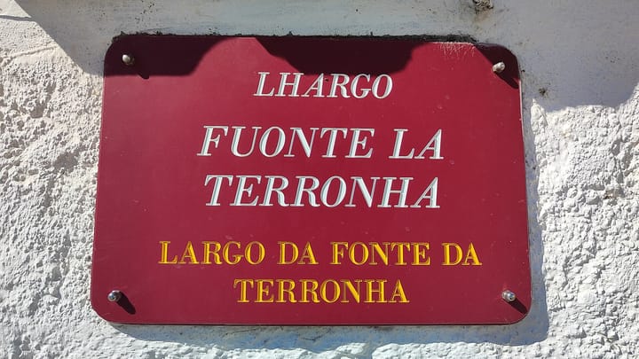 A dark red enamel street plate on a whitewashed wall reading Lhargo Fuonte la Terronha in Mirandese above Largo da Fonte da Terronha in Portuguese.