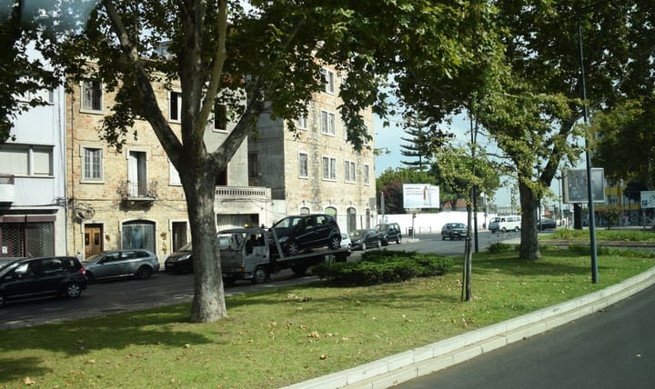 A flatbed tow truck carrying a dark hatchback along a tree-lined street in Lisbon