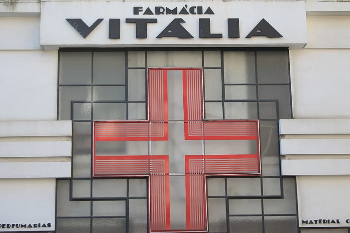 A red and white Art Deco pharmacy cross set in frosted glass under the sign of Farmácia Vitália in Porto