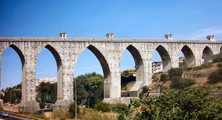 The Aqueduto das Aguas Livres in Lisbon, a long stone aqueduct on tall pointed arches crossing a valley under a clear blue sky, with apartment blocks and trees below it.