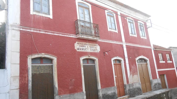 A closed village shop in Cercal, Portugal: a red painted building with the old sign Mercearia e Fazendas, Vinhos e Tabacos, Manuel Costa on the wall and the shop doors shuttered.
