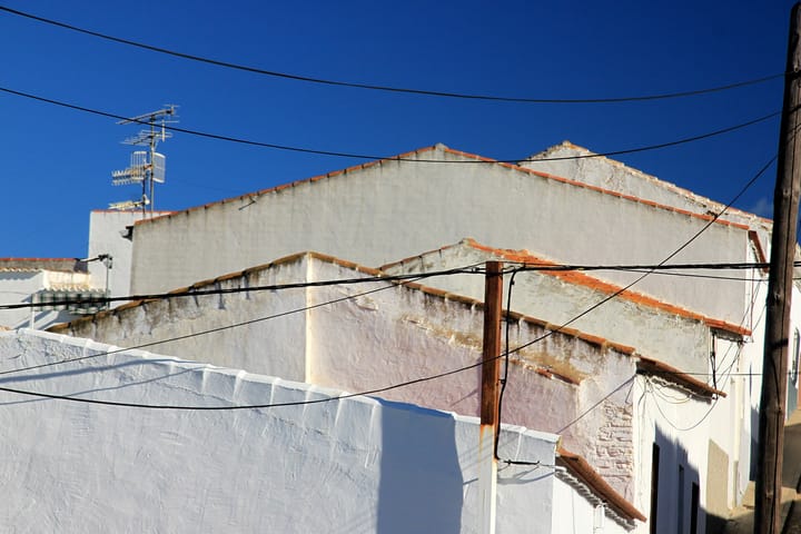 Whitewashed gable walls and terracotta tiled roofs of houses in a Portuguese village against a deep blue sky, with overhead cables and a television aerial.
