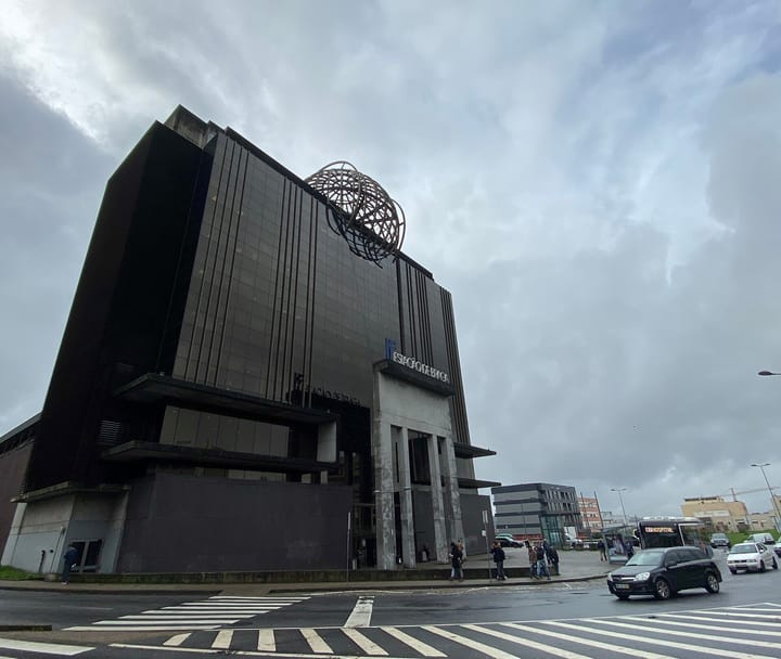 The dark glass front of Braga railway station under a grey sky, with the steel globe sculpture on its roof, the Estacao de Braga sign over the entrance and a bus waiting outside