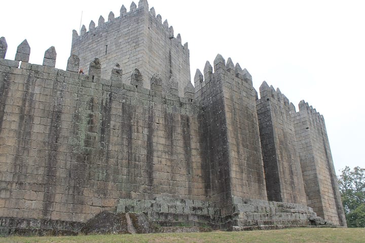 The granite keep and crenellated curtain wall of the Castelo de Guimarães seen from below on the grass, with a visitor standing on the wall walk between the merlons.