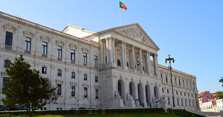 The Palacio de Sao Bento in Lisbon, seat of the Portuguese parliament, its white colonnaded portico and pediment under a clear blue sky with the national flag flying above.