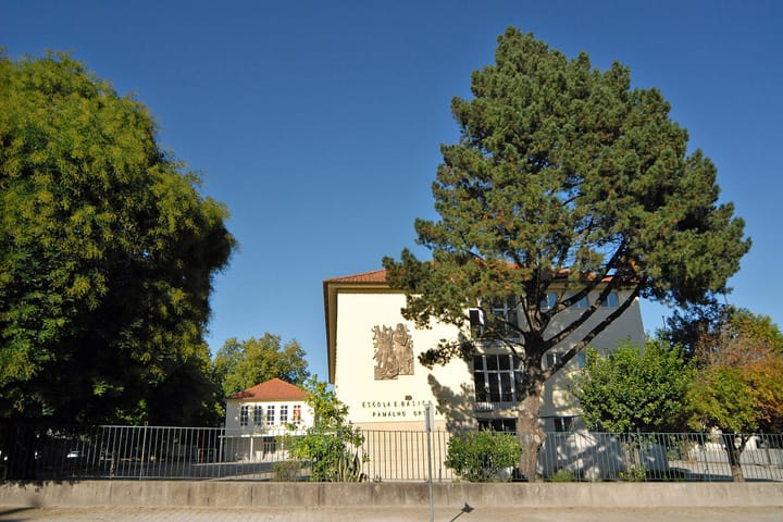 A Portuguese basic school building behind railings and pine trees, with Escola E. Basico 2,3 Ramalho Ortigao carved into the pale facade beneath a stone relief