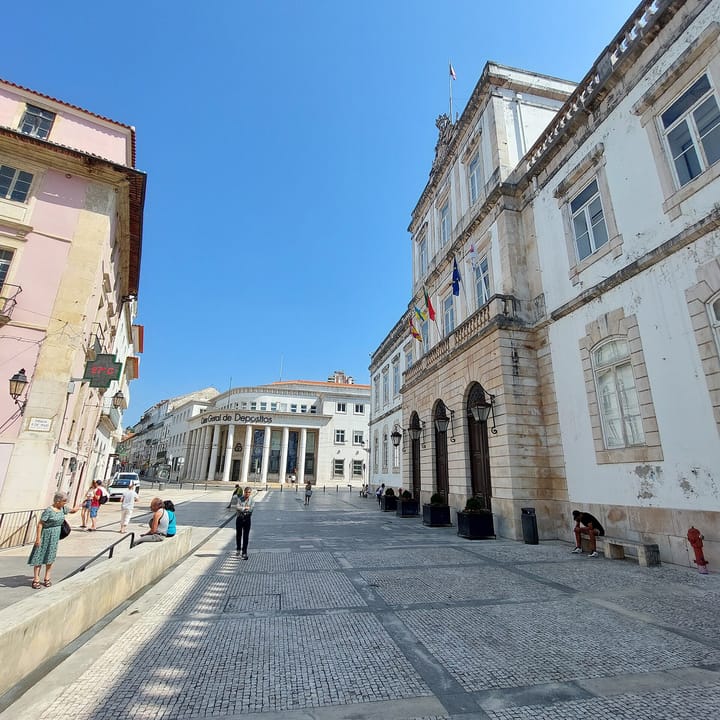 The Câmara Municipal de Coimbra on the Praça 8 de Maio, flying the Portuguese, European and municipal flags, with people crossing the square.
